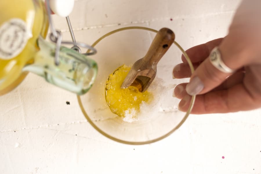 A woman's hands pour golden oil from a glass bottle into a glass bowl containing sea salts and wooden spoon.